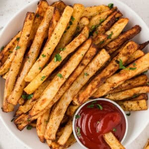 A white plate topped with oven baked French fries with ramekin of ketchup.