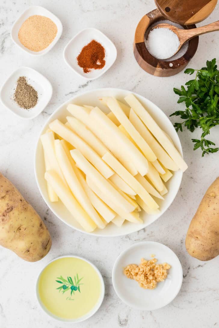 Ingredients on a light surface, including sliced potatoes, spices, oil and parsley.