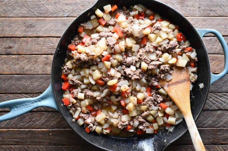 A cast iron skillet with light blue handles, filed with cubed potatoes, ground beef and red bell pepper.