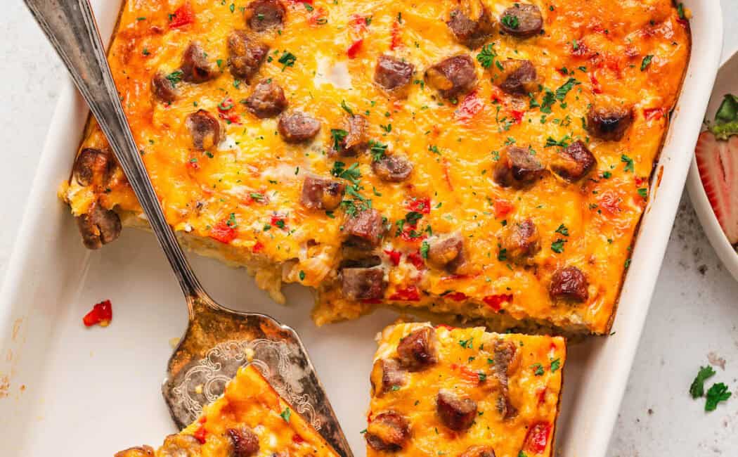 A white rectangular baking dish filled with breakfast casserole with sausage, with a few pieces removed or in the process of being removed.