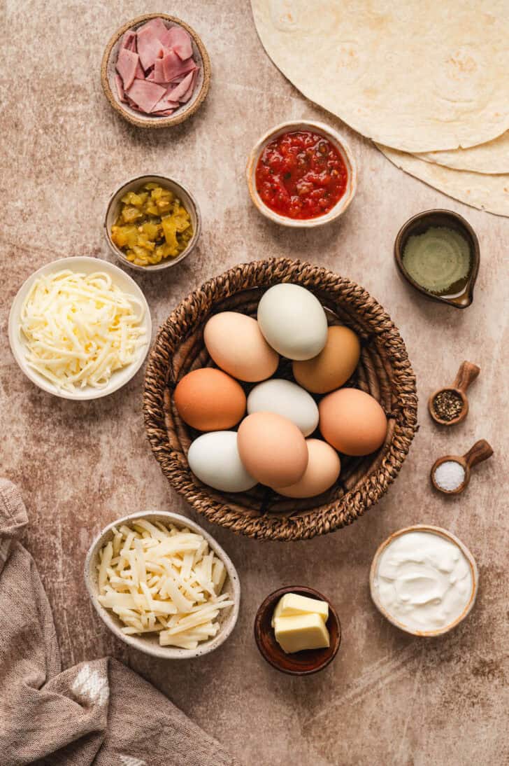 Ingredients on a rustic light brown surface, including eggs, shredded cheese, hash browns, salsa, green chiles, sour cream, butter and spices.