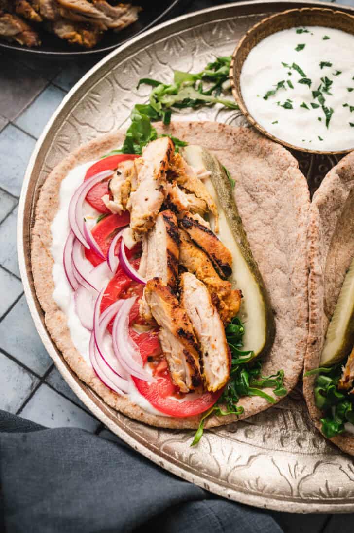 A chicken shawarma recipe presented on a plate on top of pita, with a side of yogurt sauce.