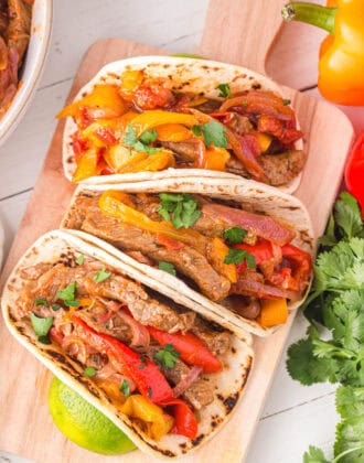 Three Instant Pot Steak Fajitas in flour tortillas, on a wooden cutting board.