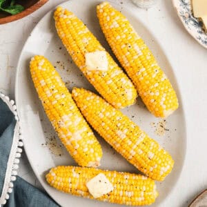 A white oval platter filled with boiled corn on the cob topped with pats of butter.