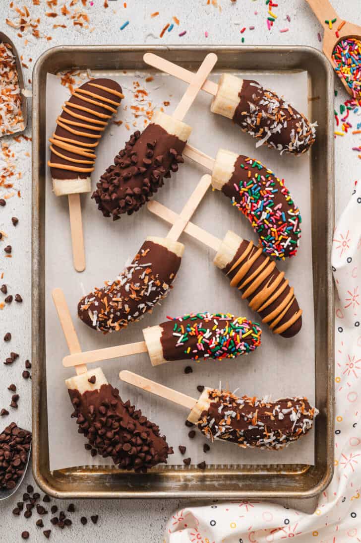 Frozen chocolate covered bananas with various toppings on a parchment-lined baking pan.