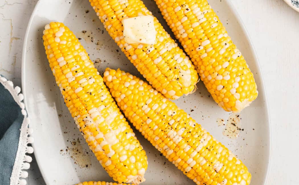 A white oval platter filled with boiled corn on the cob topped with pats of butter.