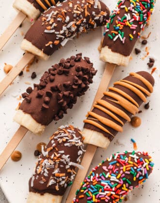 Chocolate covered bananas with various toppings on a light surface.