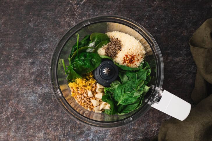 A food processor filled with herbs, cheese, nuts, lemon zest, garlic and spices.
