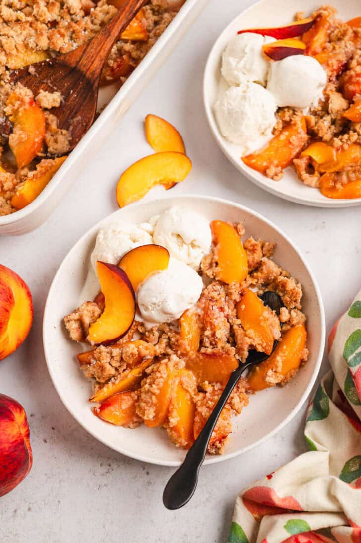 A white bowl filled with fresh peach cobbler topped with vanilla ice cream.