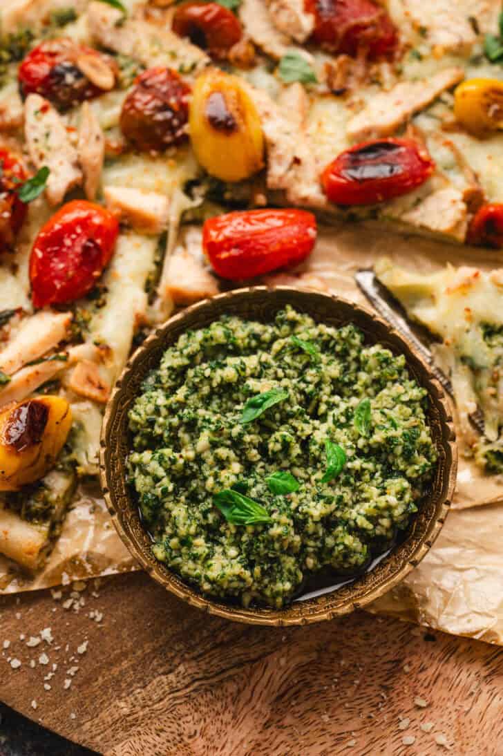 A bowl of a homemade pesto sauce recipe in front of a cut pizza with blistered tomatoes on it.