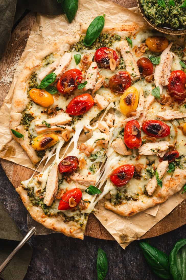 A pesto pizza topped with blistered tomatoes, with a serving spatula taking a slice out.