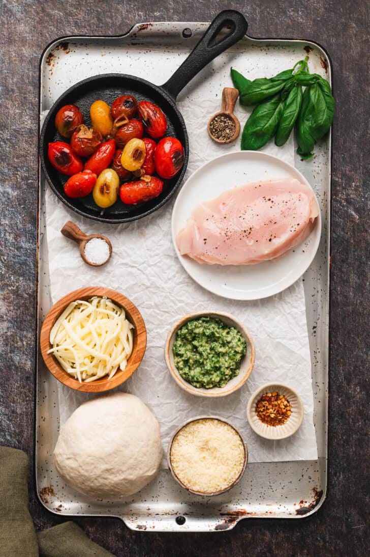 Ingredients on a light speckled platter, including poultry, tomatoes, cheese, an herb mixture, basil, pizza dough and spices.