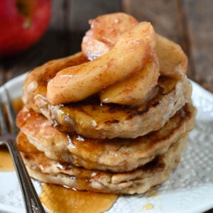 A stack of three apple pancakes topped with sauteed apples and maple syrup on a textured white plate.