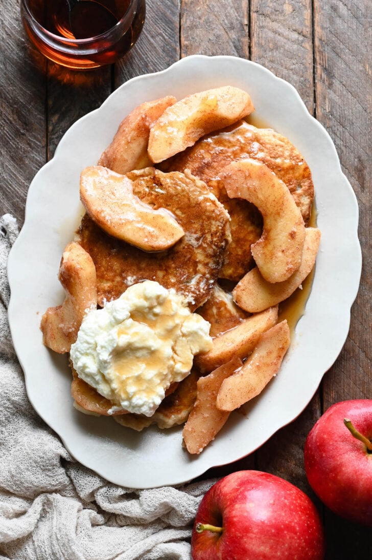 A white scalloped platter topped with an apple pancakes recipe, sauteed apples, whipped cream and maple syrup.