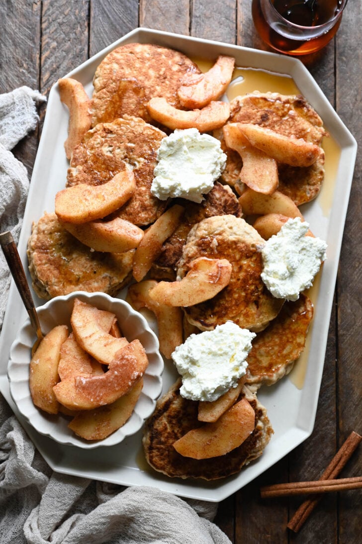 A geometric white platter filled with apple pancakes topped with sauteed apples, whipped cream and maple syrup.