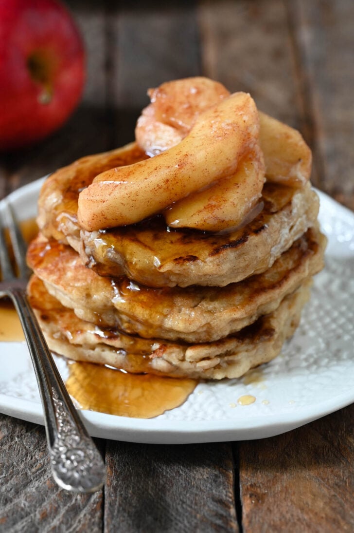 A stack of three apple pancakes topped with sauteed apples and maple syrup on a textured white plate.