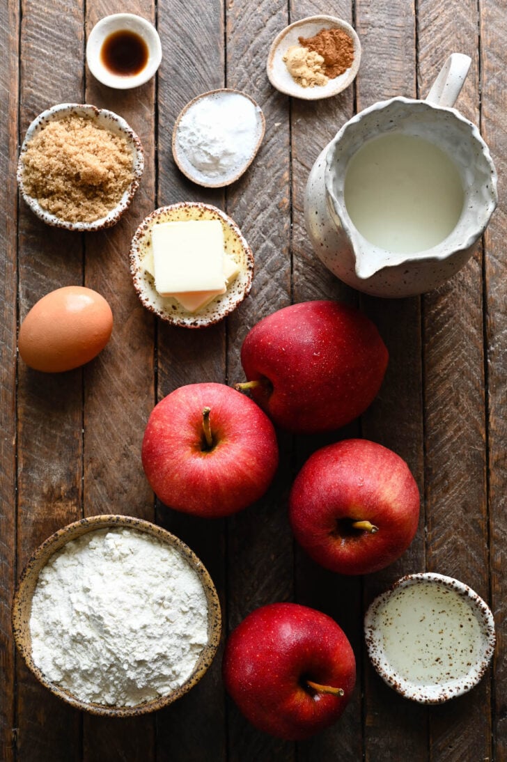 Ingrediens on a wooden surface, including red apples, flour, brown sugar, butter, buttermilk, spices, oil and an egg.