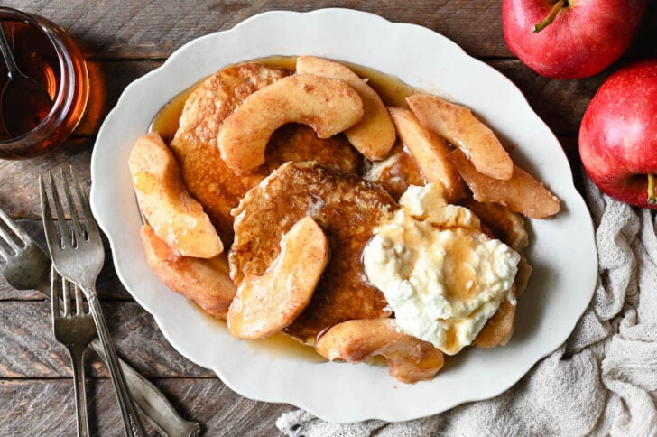 A white scalloped platter topped with apple cinnamon pancakes, sauteed apples, whipped cream and maple syrup.