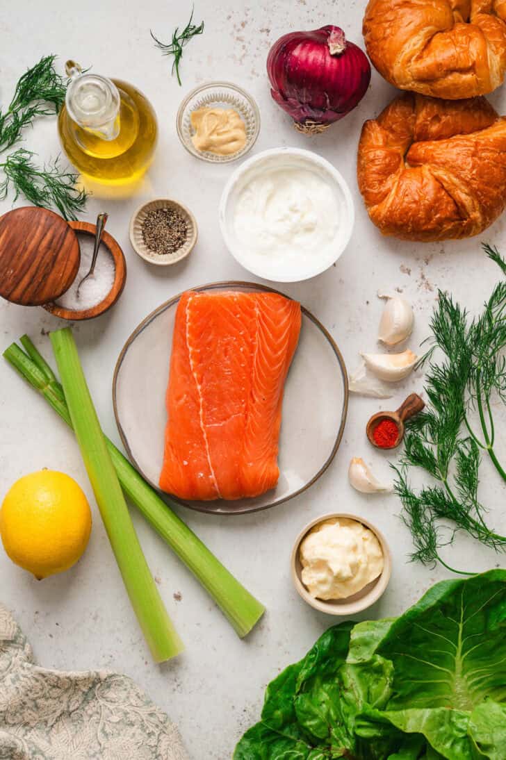 Ingredients on a white surface, including raw fish, mayonnaise, yogurt, celery, herbs, lemon, spices, garlic, onion and croissants.