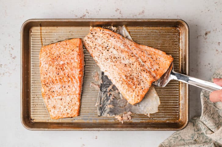 Two pieces of cooked orange fish on a rimmed baking pan, with a spatula removing one of the fish fillets from the skin.