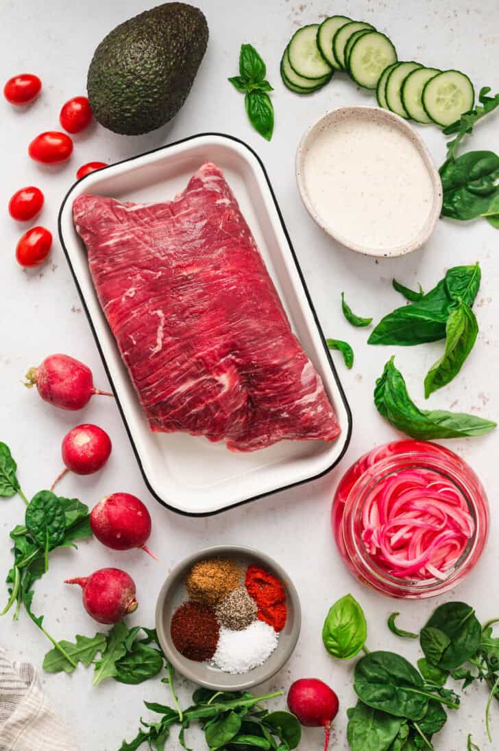 Ingredients on a white surface, including raw beef, greens, pickled red onions, avocado, cucumber, spices, radishes and creamy dressing.