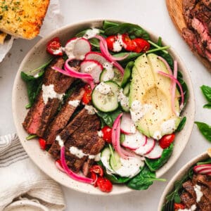 A dinner scene with bowls of a steak salad recipe alongside sliced steak, pickled red onions and garlic bread.