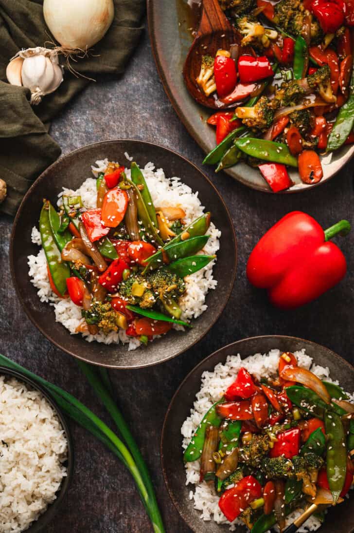 Bowls filled with veggie stir fry over white rice on a dark surface.