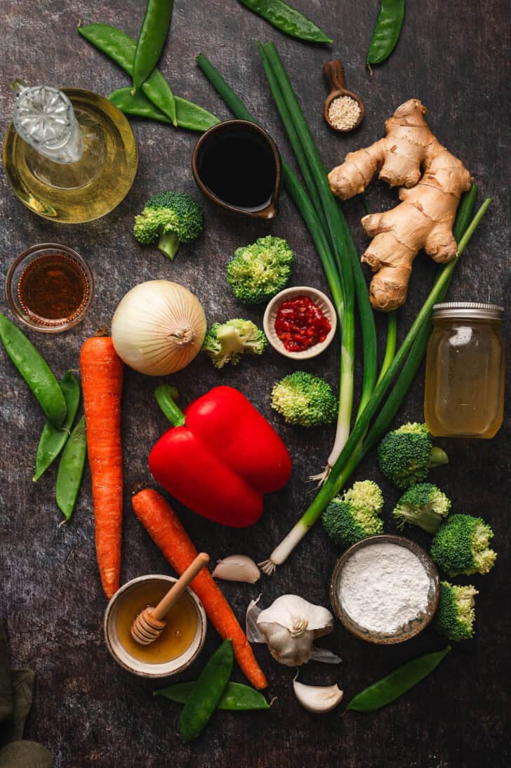 Ingredients on a dark surface, including broccoli, red pepper, carrots, snow pea pods, green onions, garlic, honey broth, ginger, soy sauce and spices.