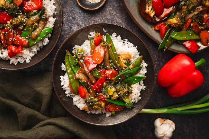 Bowls filled with vegetarian stir fry over white rice on a dark surface.