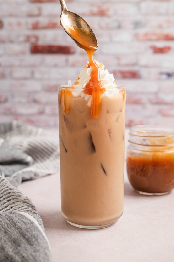 Caramel sauce being spooned over a coffeehouse drink topped with whipped cream.