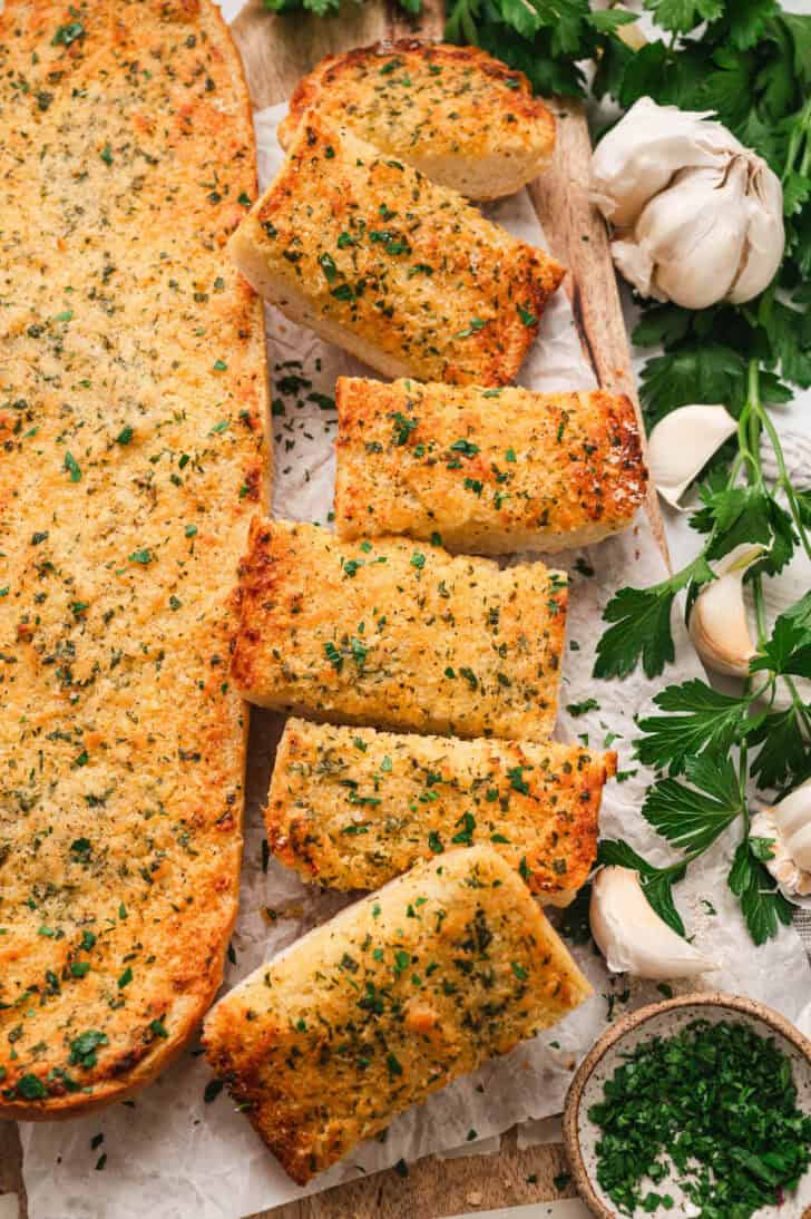 A loaf of a homemade garlic bread recipe on a wooden cutting board on a light surface.