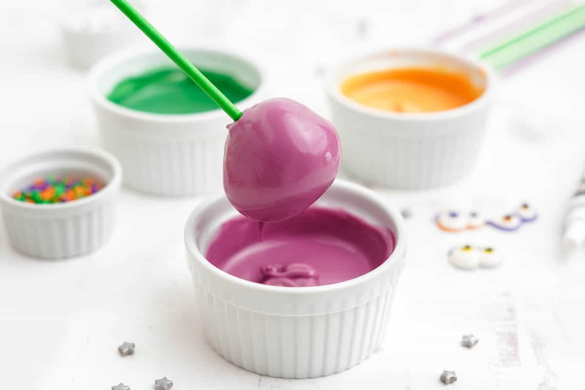 A green stick dipping a donut hole into purple candy coating.
