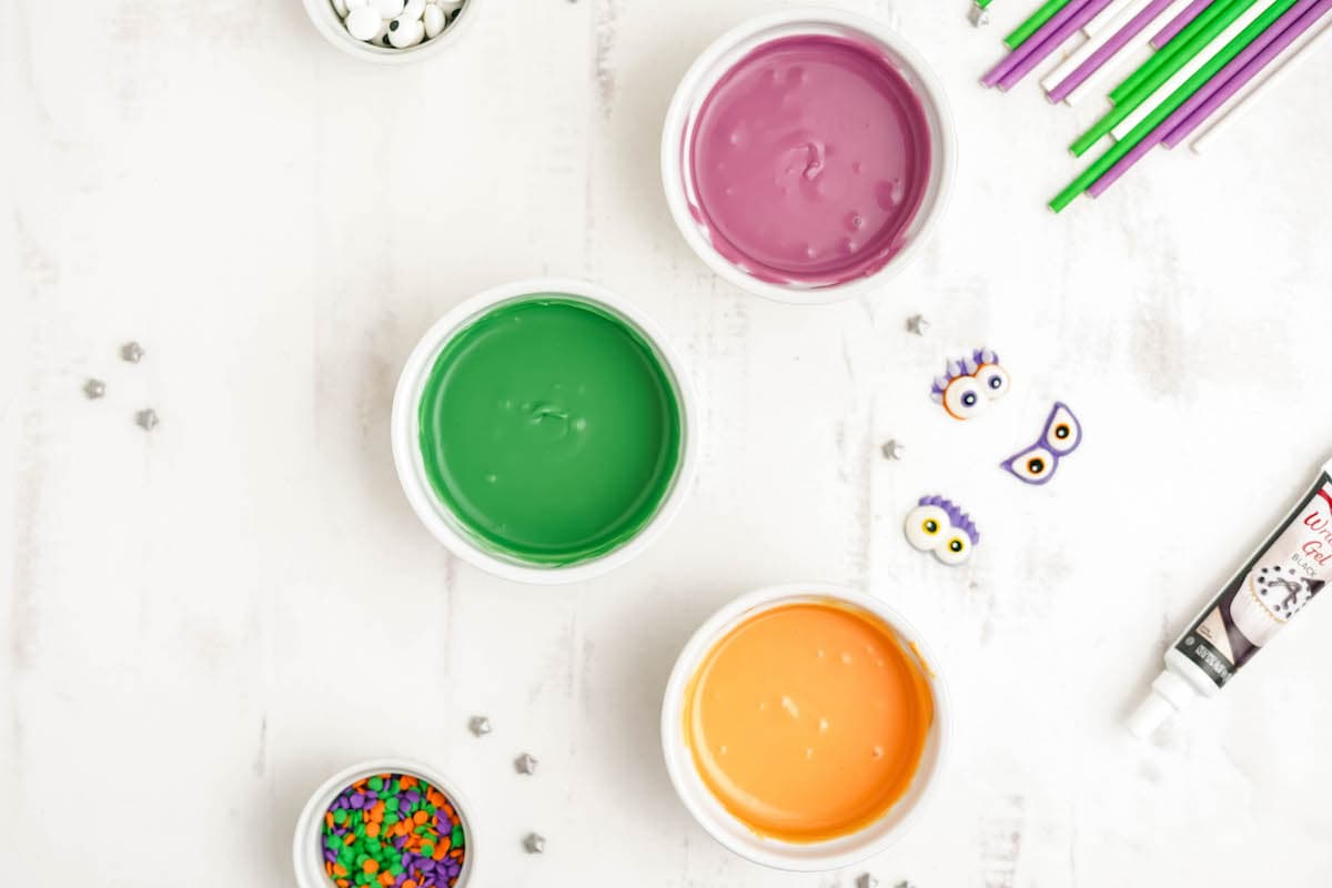 Three ramekins of melted candy melts in purple, green and orange on a white surface.