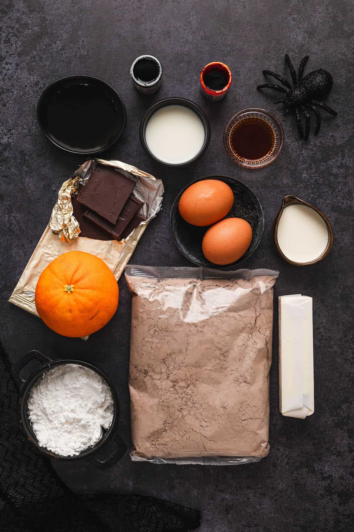 Ingredients on a dark surface, including baking mix, chocolate, and eggs, orange and butter.