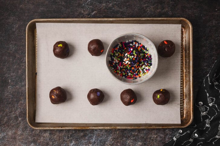 Chocolate cookie dough balls on a rimmed baking pan with a bowl of sprinkles.