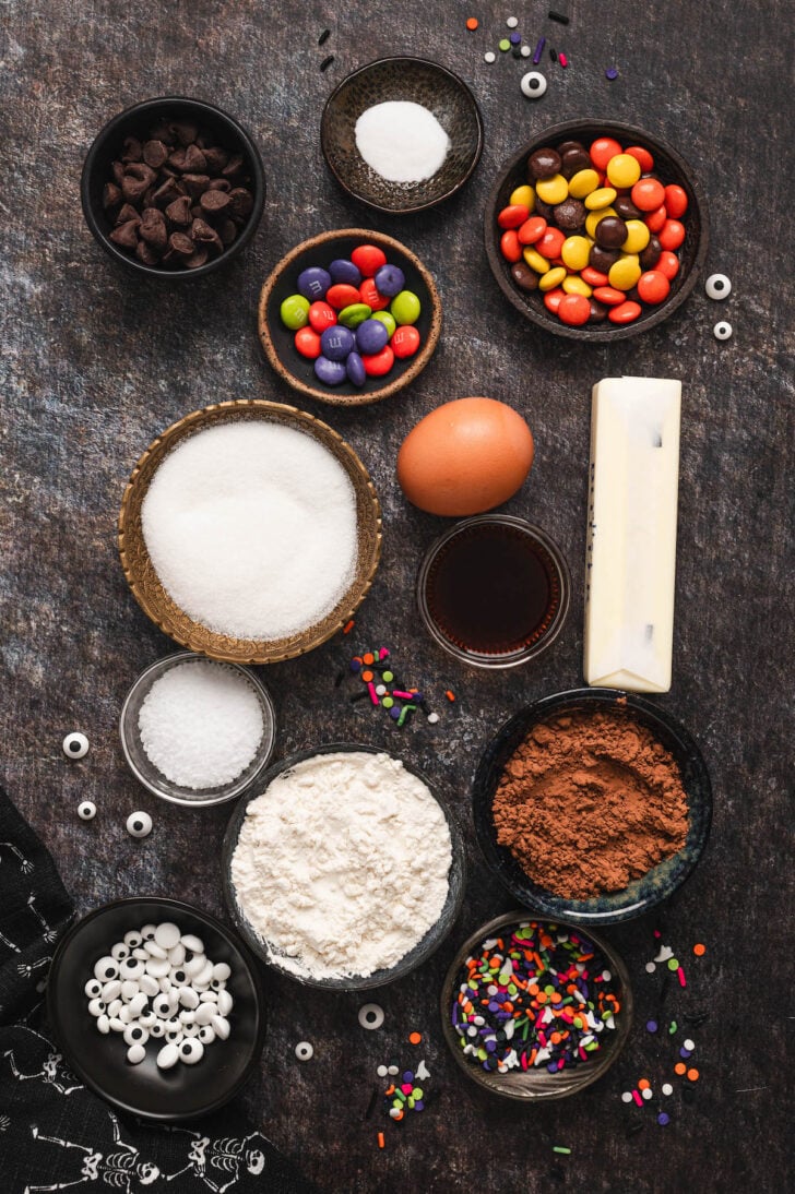 Baking ingredients on a dark surface including flour, sugar, butter, cocoa powder, candy, chocolate chips, candy eyeballs and sprinkles.