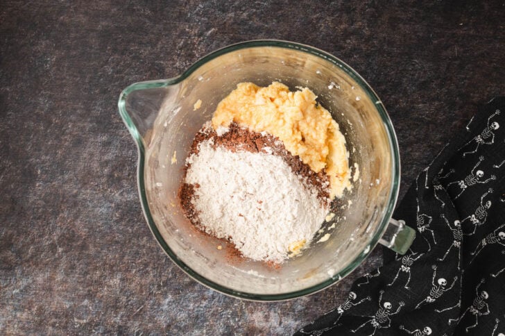 A glass bowl of a stand mixer filled with creamed butter, sugar and egg, and flour and cocoa powder.