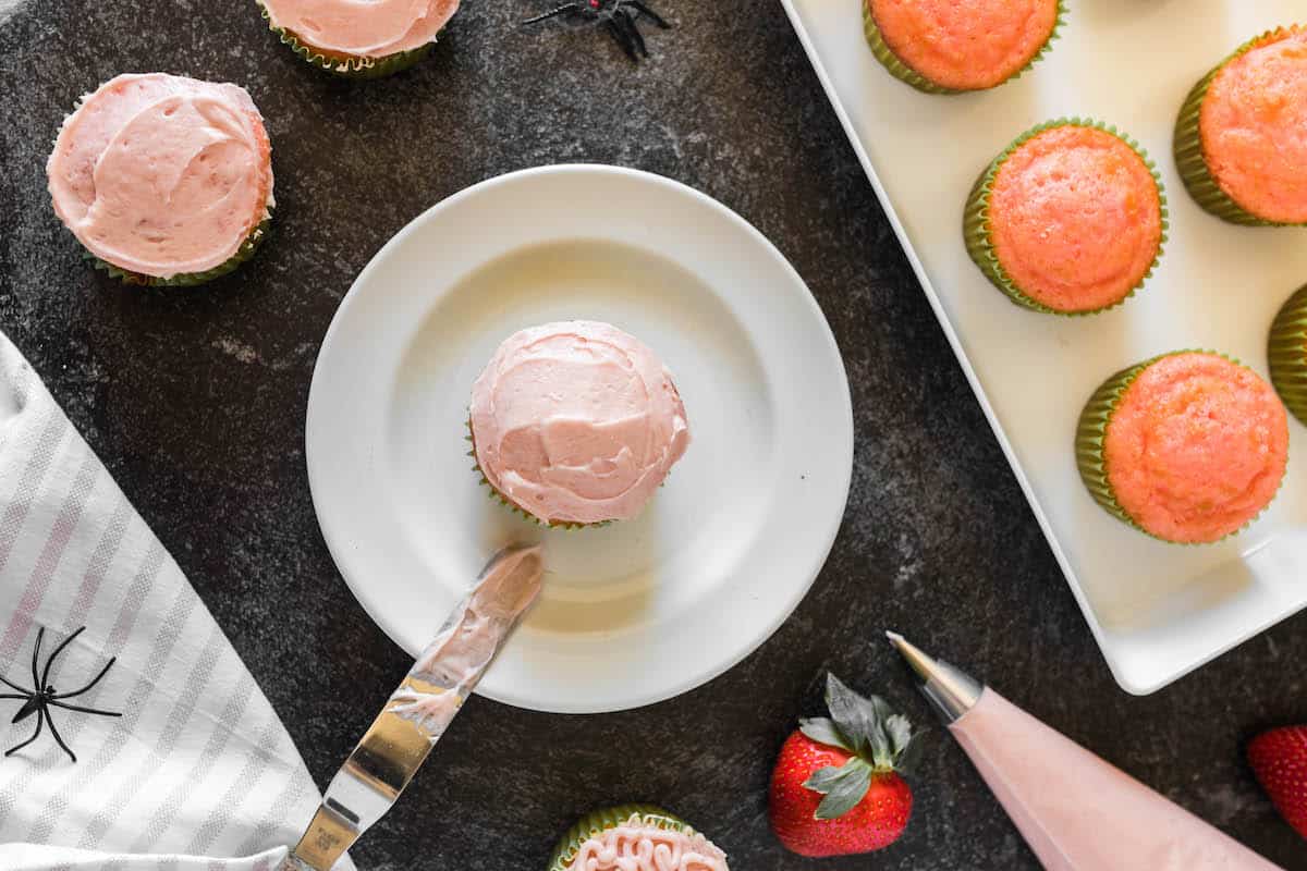 A cupcake being frosted with pink frosting on a white plate.