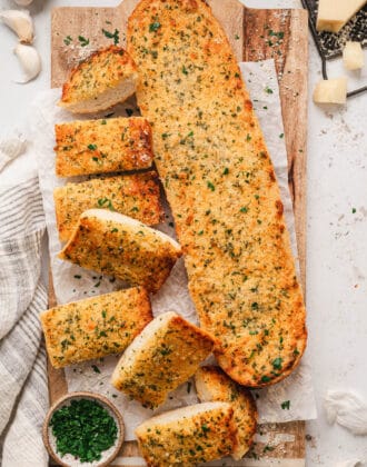 A loaf of homemade garlic bread on a wooden cutting board on a light surface.