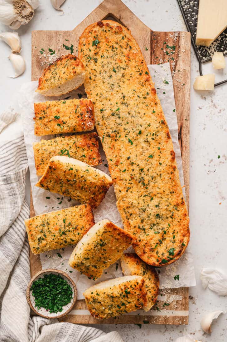 A loaf of homemade garlic bread on a wooden cutting board on a light surface.