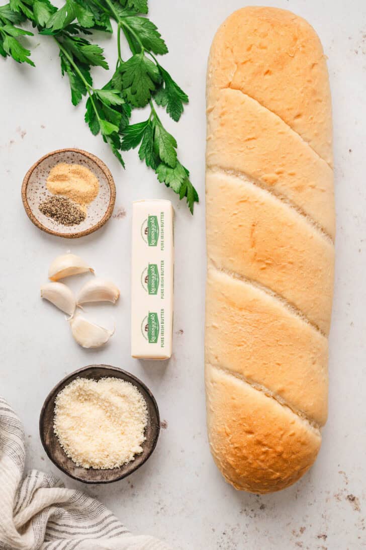 Ingredients on a white surface including a loaf, spices, Parmesan cheese, garlic cloves and a stick of butter.