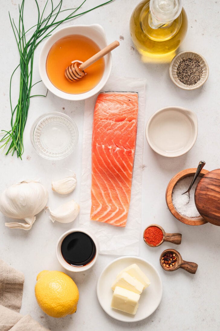 Ingredients on a white surface, including a piece of raw pink fish, garlic, soy sauce, spices, lemon, butter and oil.