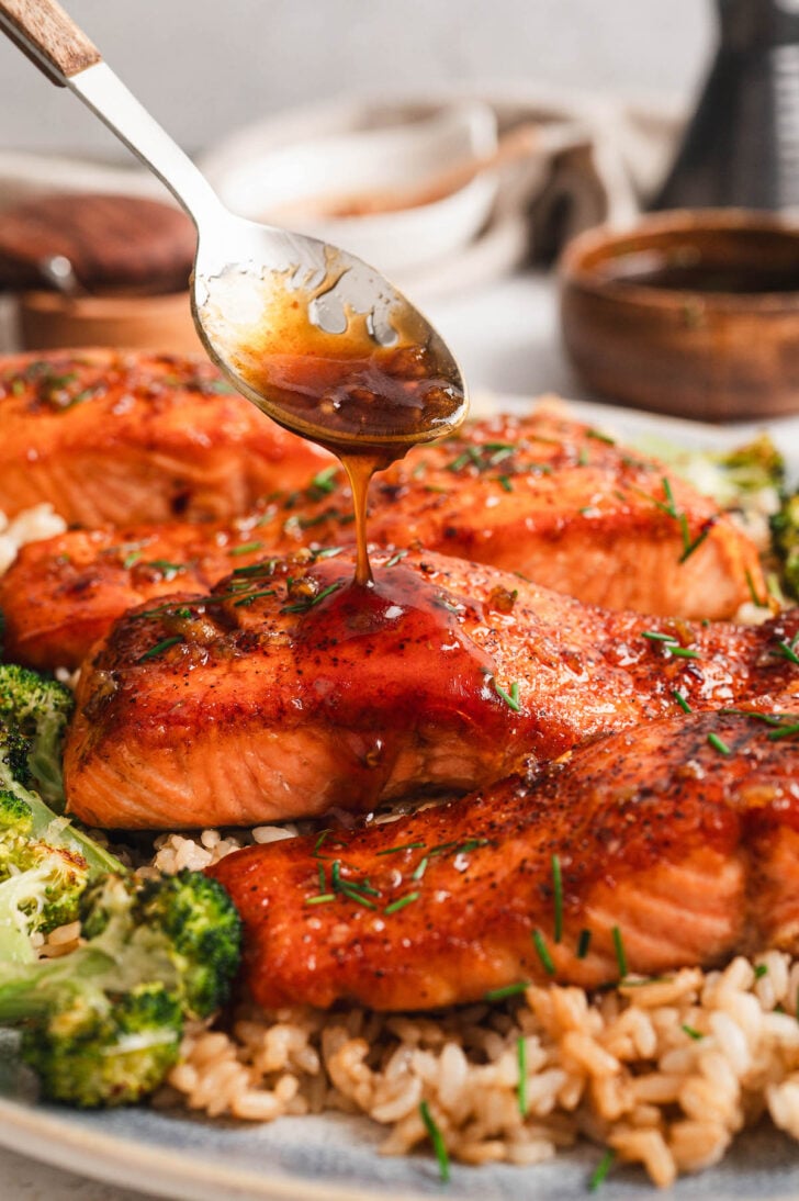 Four pieces of a honey glazed salmon recipe on a platter, served over rice and broccoli, being drizzled with glaze.