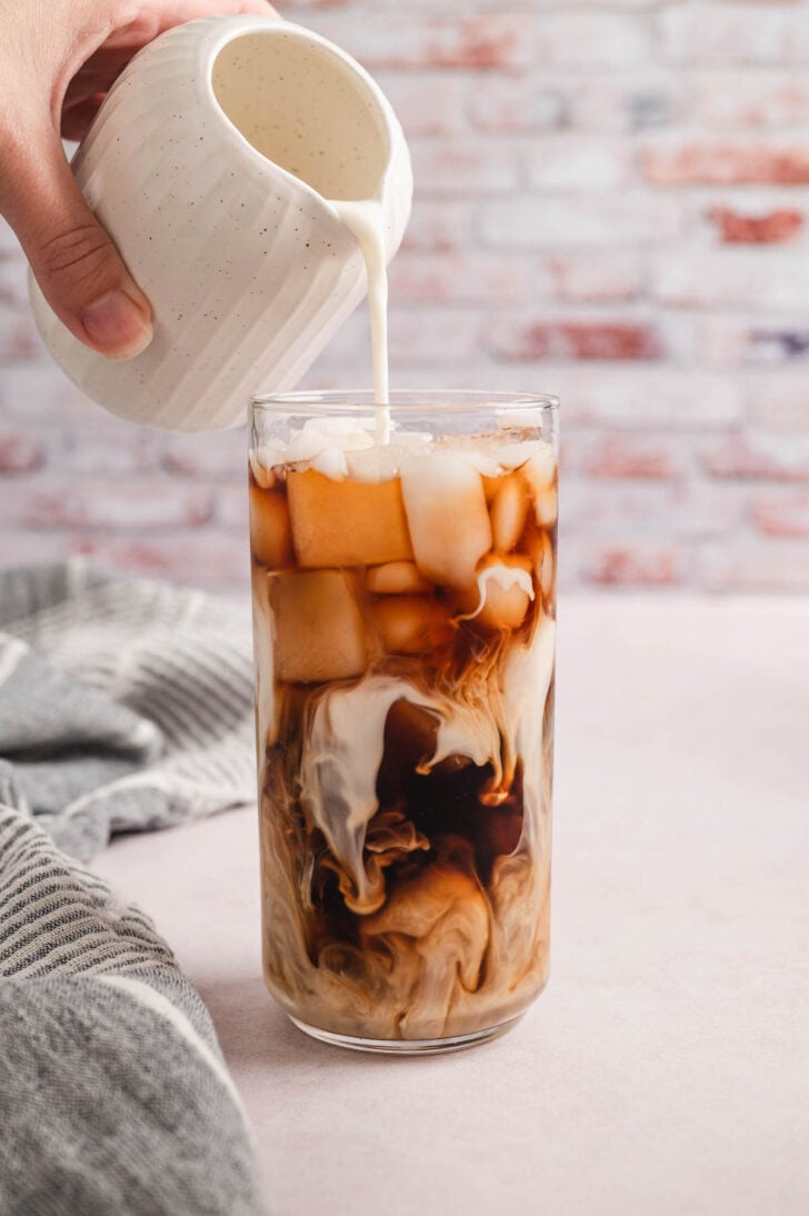 A hand pouring milk from a pitcher into a tall glass of iced cold brew.