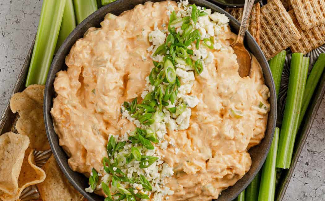 A bowl of buffalo chicken dip on a platter with cut veggies, crackers and tortilla chips.