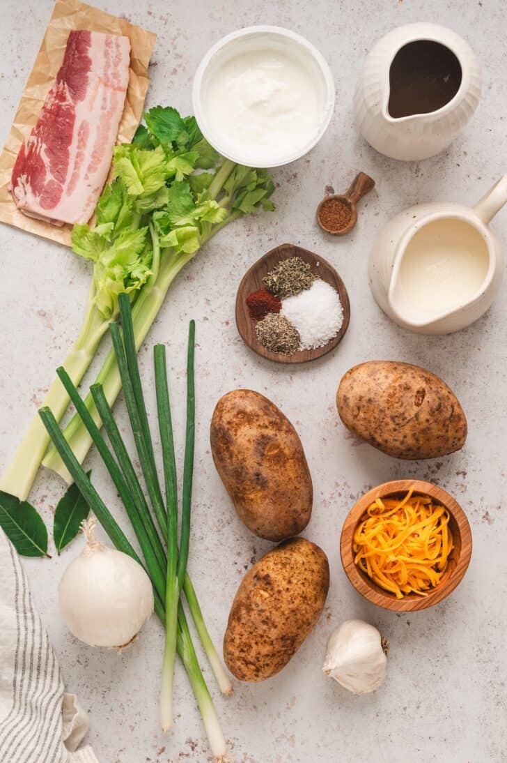 Ingredients on a light surface, including potatoes, onions, celery, bacon, spices, cheese, garlic, yogurt, half and half and chicken broth.