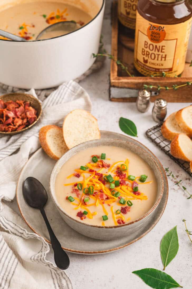 A bowl of easy potato soup recipe topped with bacon, green onions and shredded cheese, with jars of Zoup! bone broth in the background.