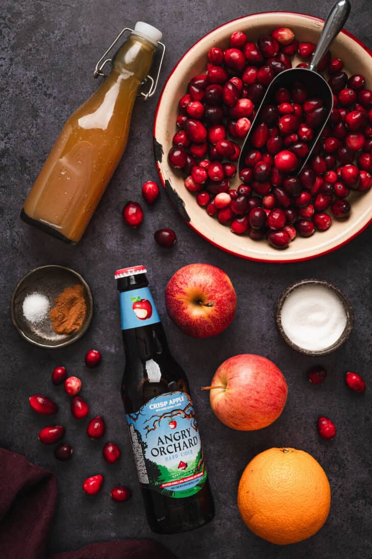 Ingredients on a dark surface, including berries, apples, sugar, spices, orange and a bottle of hard apple cider.