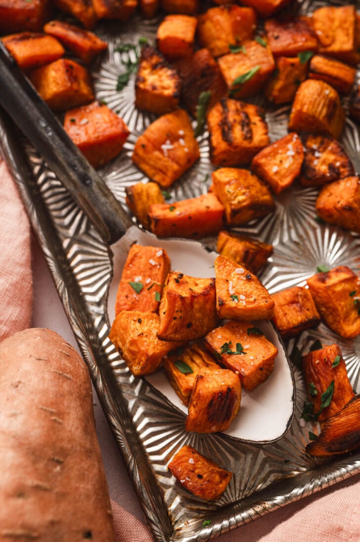 Oven roasted sweet potatoes on a serving spoon on a textured baking pan.