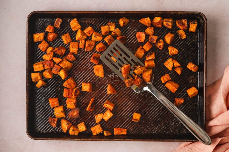 Cubed orange root vegetables being tossed with a pancake turner on a baking pan.